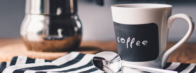 Tasse Kaffee mit Löffel und Kanne im Hintergrund.